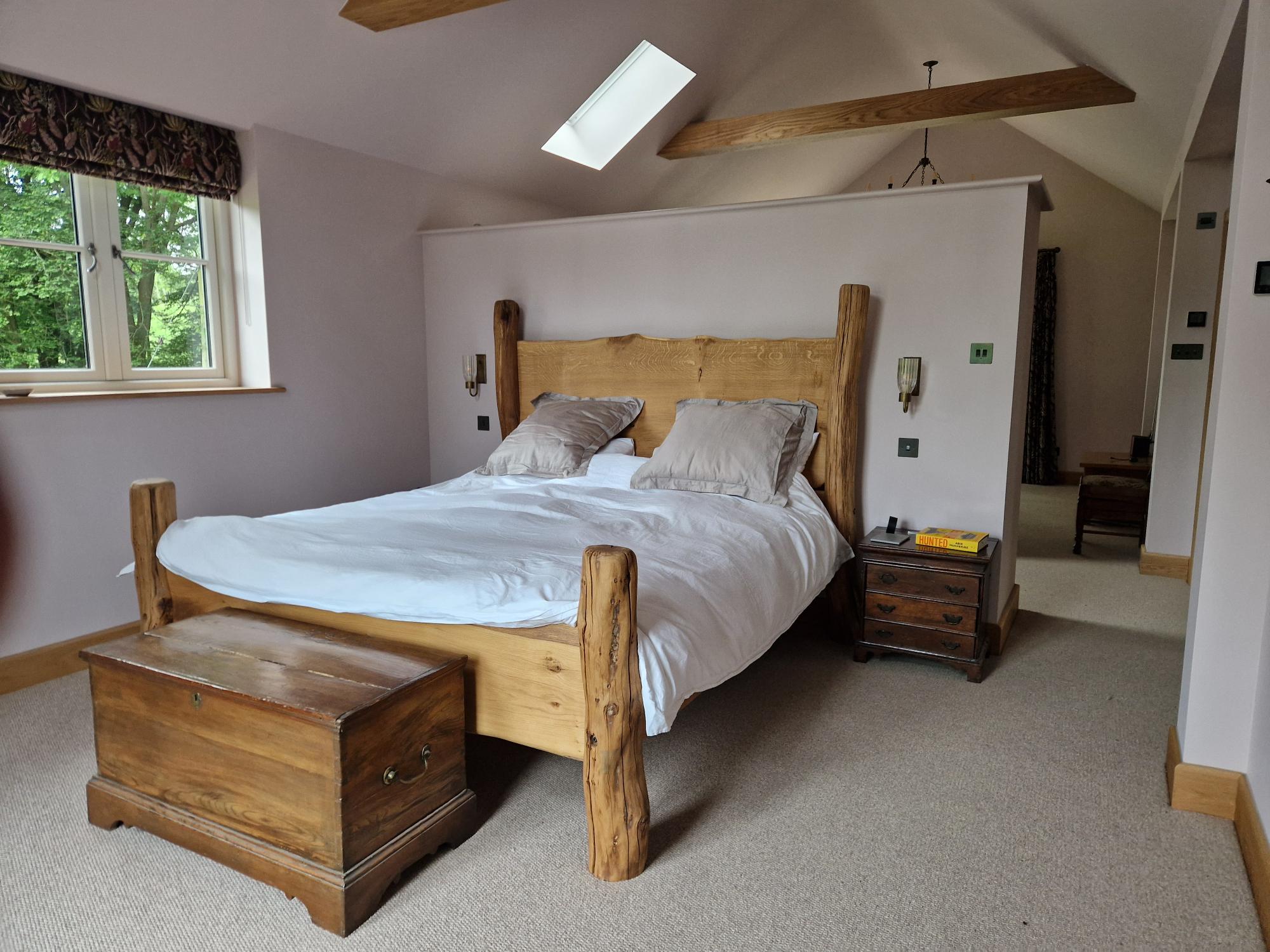 Solid oak handcrafted bed with a rustic design, set in a spacious bedroom with natural light. The bed features sturdy wooden posts and a smooth wooden headboard, complemented by a wooden chest at the foot of the bed